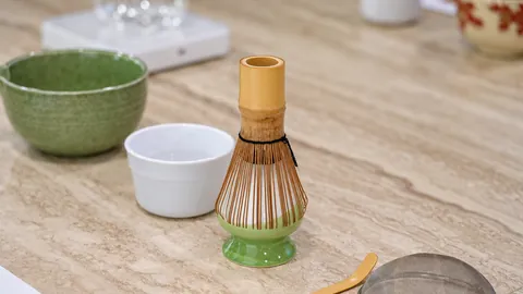A bamboo matcha whisk beside a ceramic bowl and cup